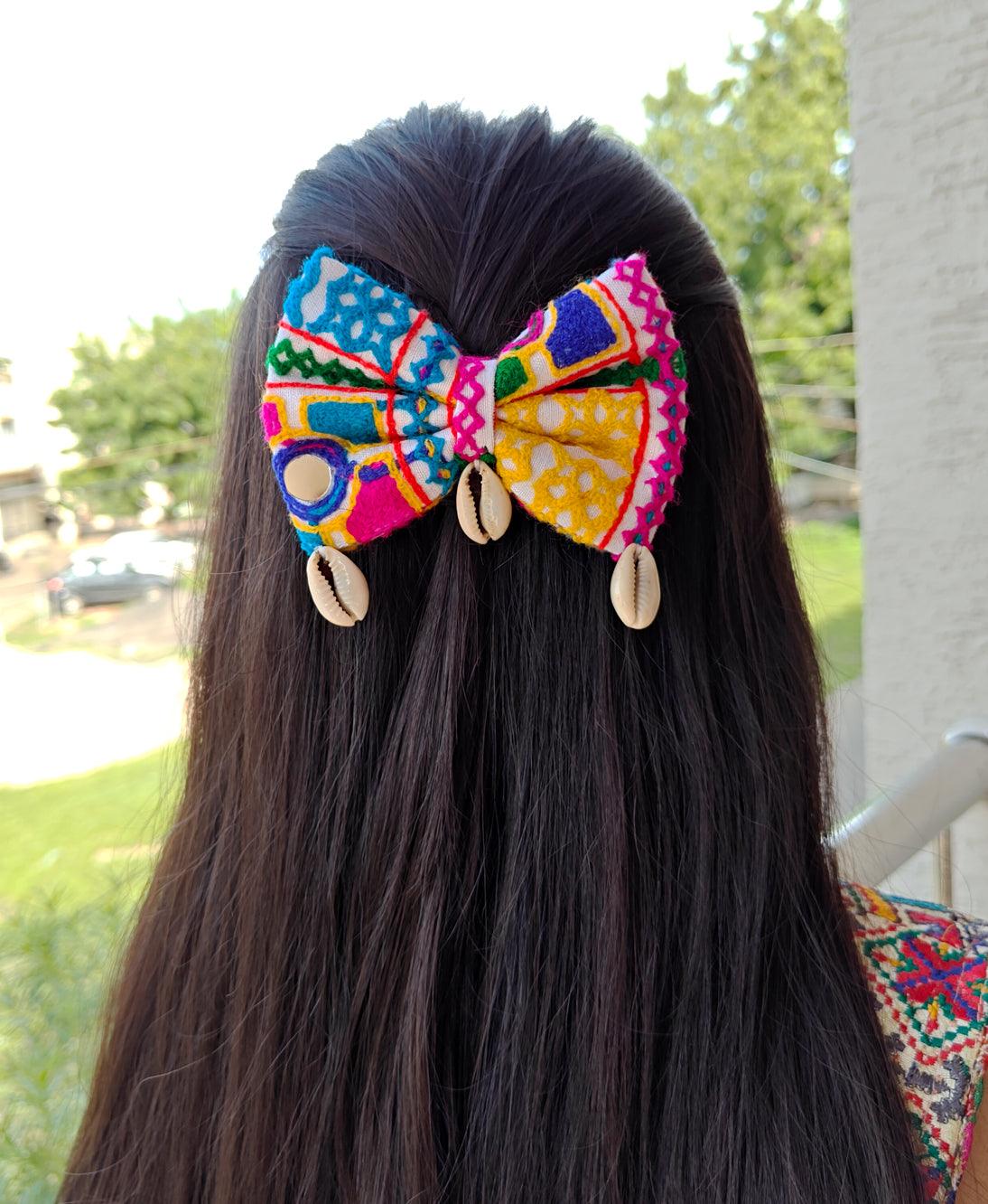 Woman with long hair wearing a vibrant Navratri-themed bow featuring colorful embroidery and cowrie shell accents, set against an outdoor background.