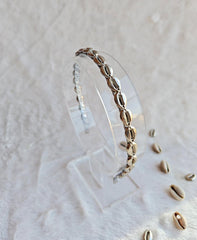 A delicate silver headband adorned with cowrie shell-shaped beads displayed on a clear stand. The bracelet features a series of metallic cowrie beads, creating a simple yet elegant accessory. The backdrop is a soft, light-colored fabric with a few scattered cowrie beads, enhancing the navratri-inspired theme of the jewelry.