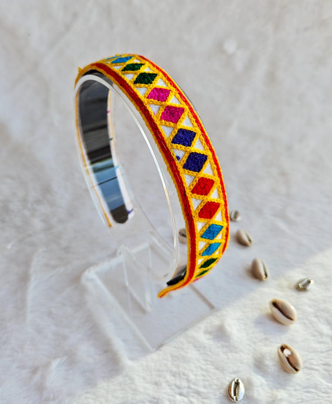 Colorful embroidered bracelet with geometric patterns displayed on a clear stand, featuring vibrant diamond shapes in red, blue, green, and yellow.