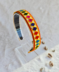 Colorful embroidered bracelet with geometric patterns displayed on a clear stand, featuring vibrant diamond shapes in red, blue, green, and yellow.