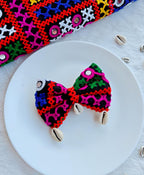 Colorful Navratri-themed bow with intricate embroidery and cowrie shell accents, displayed on a white plate with a vibrant embroidered fabric backdrop.