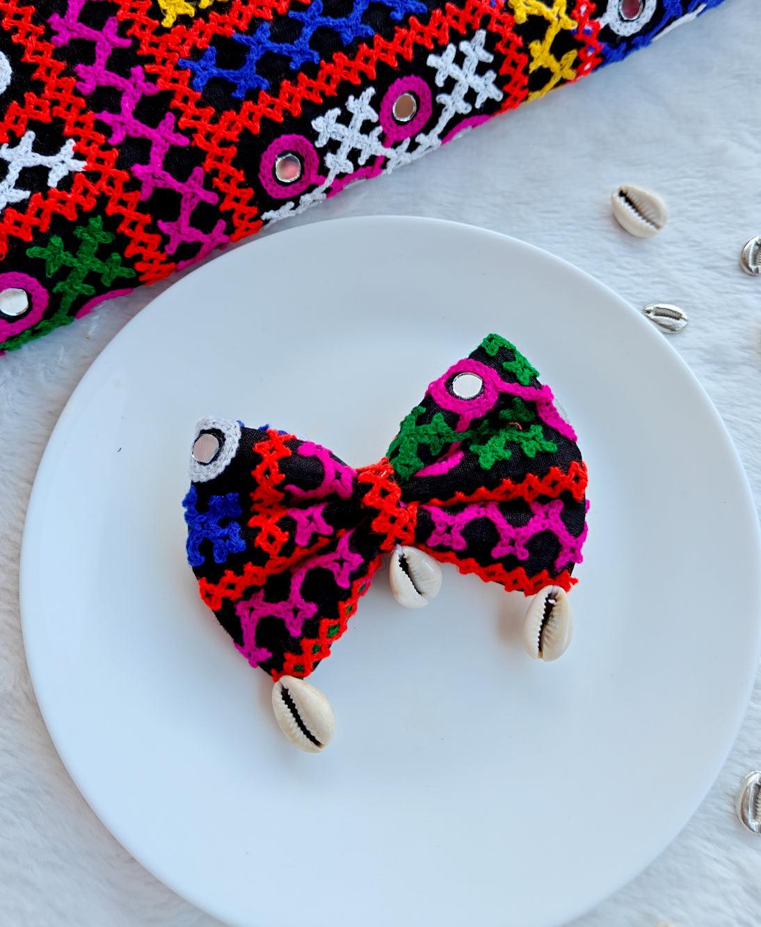 Colorful Navratri-themed bow with intricate embroidery and cowrie shell accents, displayed on a white plate with a vibrant embroidered fabric backdrop.