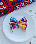 Colorful Navratri-themed bow with intricate embroidered patterns and cowrie shell accents, displayed on a white plate with a vibrant embroidered fabric backdrop.